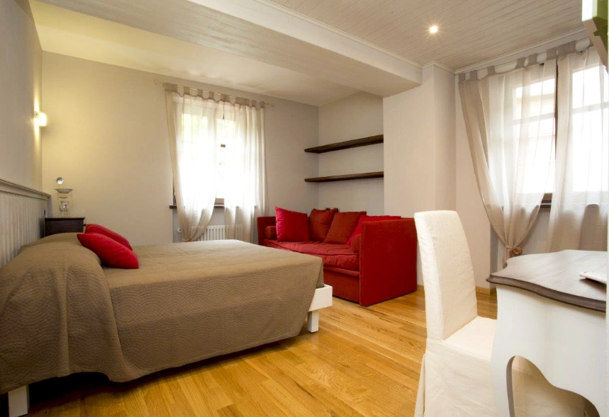 foto principale della camera Azalea con un letto matrimoniale e un Sofà di fronte ad esso, due mensole sopra il Sofà, una sedia insieme ad un tavolo e due finestre